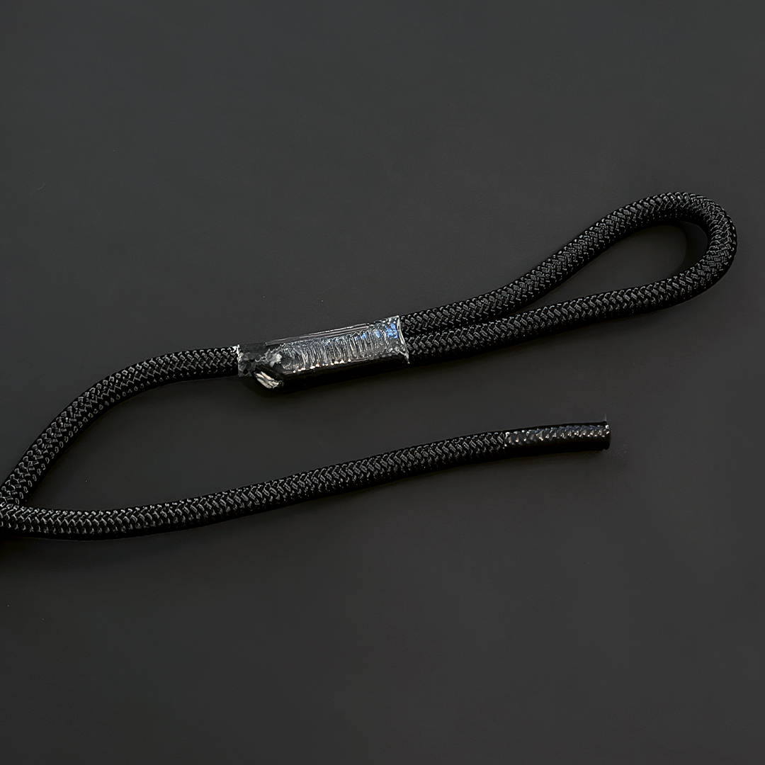 A black braided cord or strap with a silver-toned metal clasp, photographed on a dark gray background. Replacement rope / tether from Raccoon Rigs.