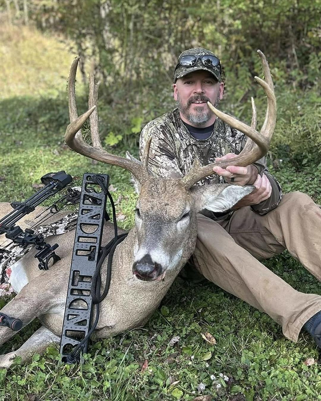 A man in camouflage clothing and a cap sits on the ground outdoors with a harvested deer in front of him, posed with a Bandit Climber by Raccoon Rigs. A compound bow is visible on the left side of the frame.