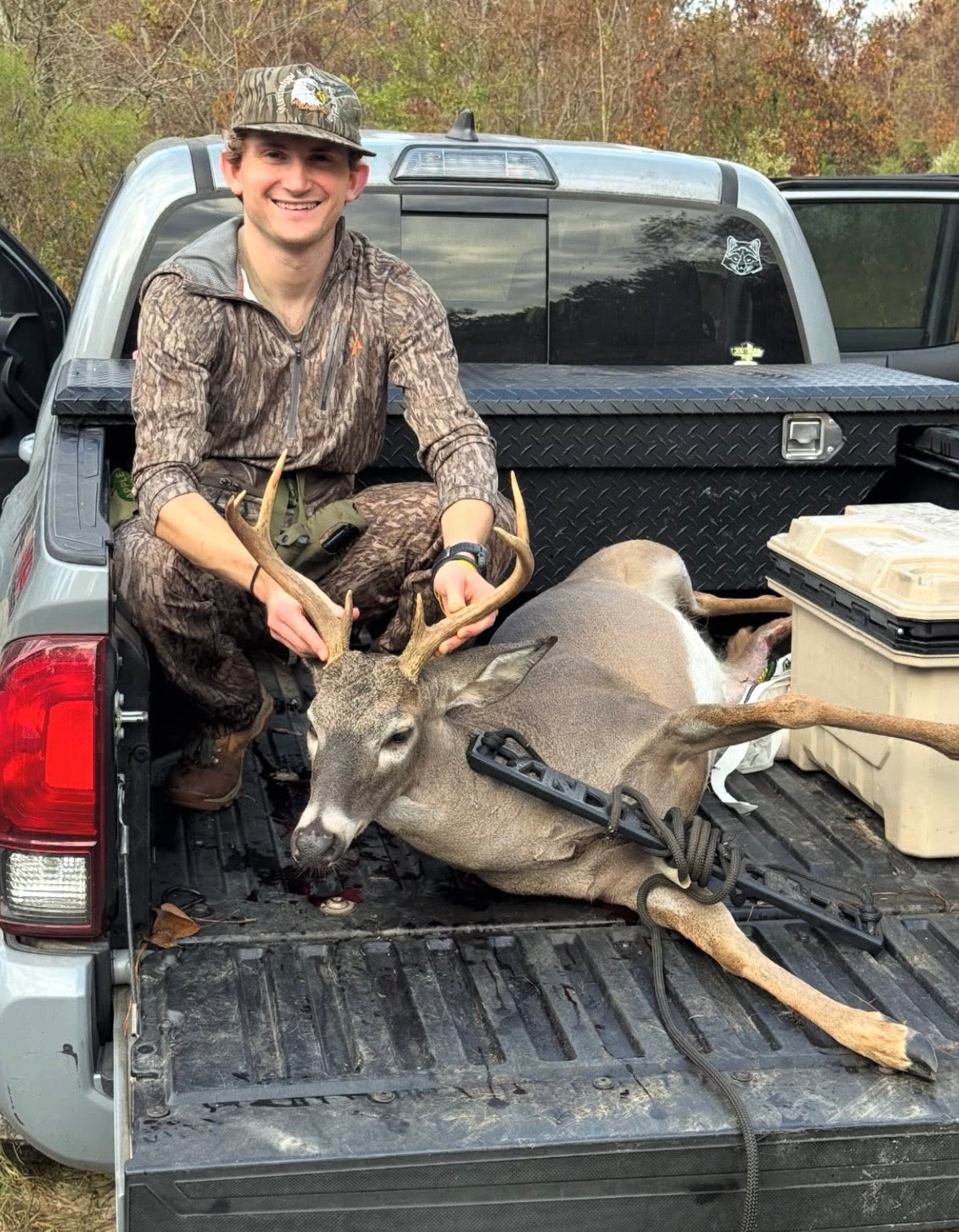 A man in camouflage clothing sits in the bed of a silver pickup truck with a harvested deer resting on the tailgate. The man is smiling at the camera, and the truck bed is lined with a black diamond-plate mat. He is using a BANDIT Climber from Raccoon Rigs.