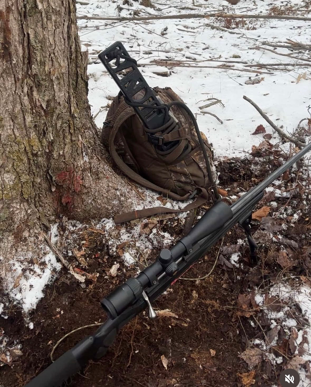A black bolt-action rifle with a scope and bipod rests on the ground next to a tree, with a brown backpack. There is a Bandit Climber from Raccoon Rigs against the tree.