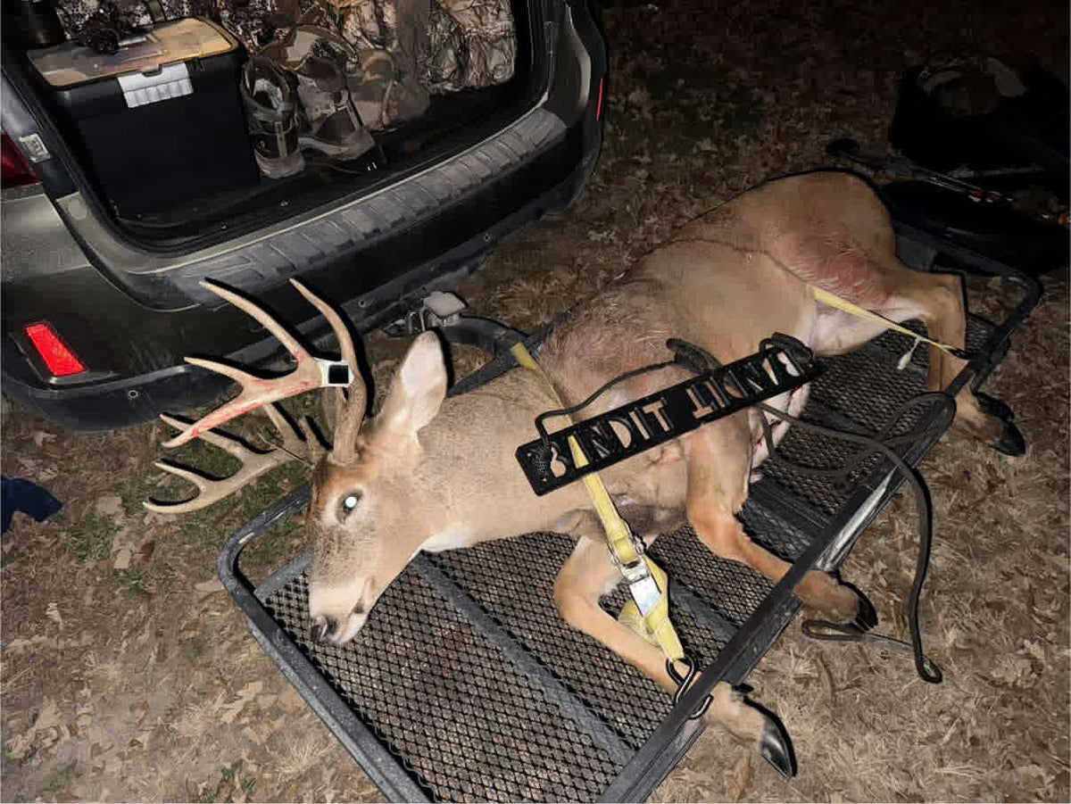 A harvested deer with antlers lies on a black metal rack trailer next to an open vehicle trunk at night, with a yellow strap and black tag attached. A Bandit Climber from Raccoon Rigs is on top of the deer.