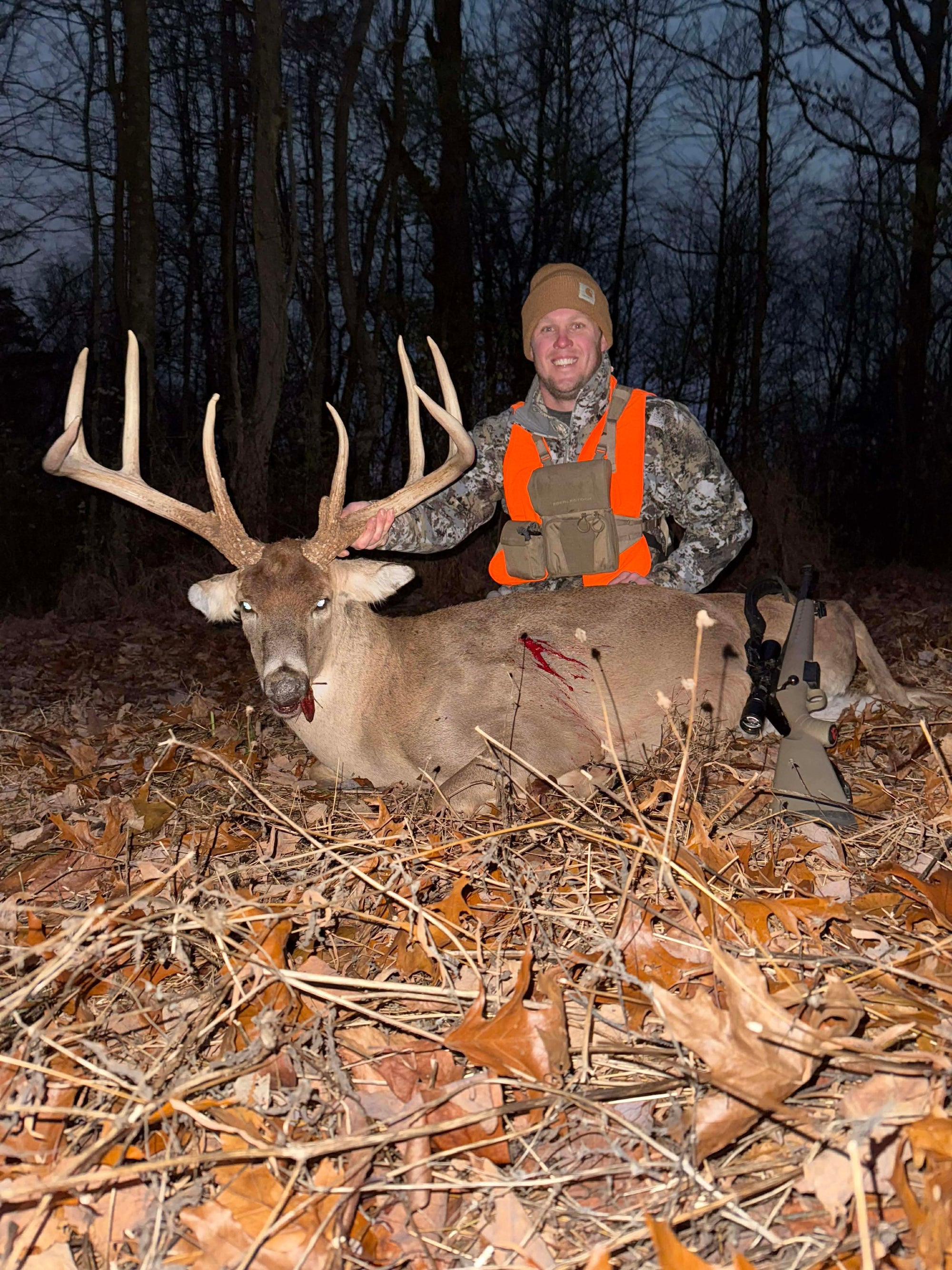A man in camouflage hunting clothing and a bright orange vest kneels behind a large buck with antlers on a bed of dry leaves in a wooded outdoor setting. He is using a Bandit Climber from Raccoon Rigs.