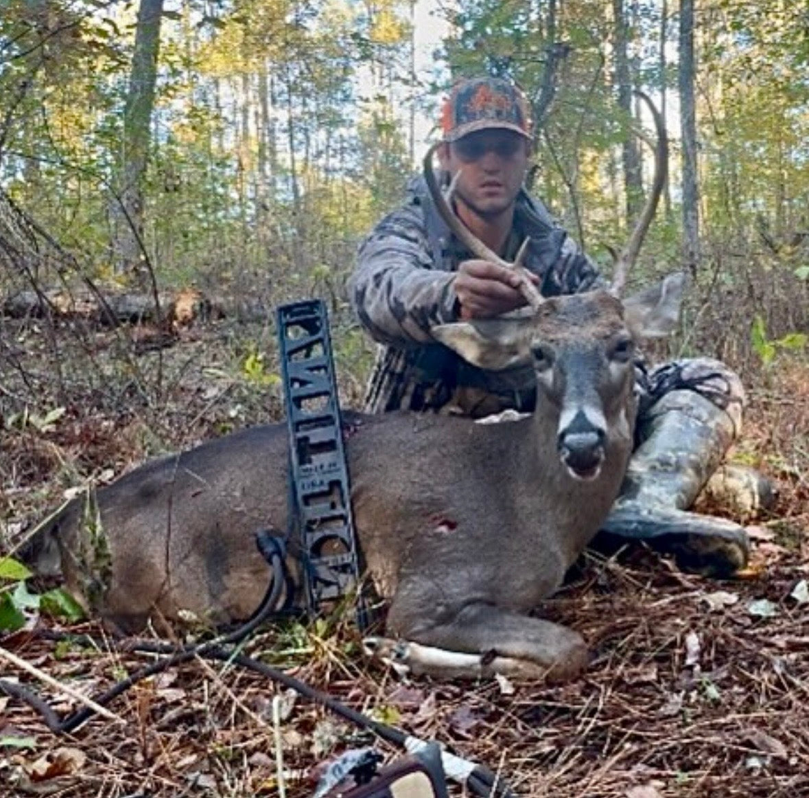 A man in camouflage hunting clothing kneels beside a harvested deer in a wooded outdoor setting, with a black compound bow resting on the deer. He is using a BANDIT Climber from Raccoon Rigs.