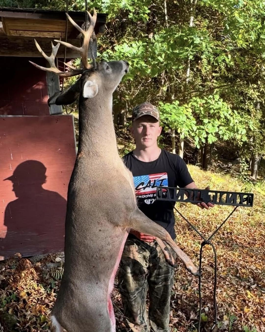 A man standing outdoors in a wooded area next to a harvested deer with large antlers, holding a black metal rack. He is wearing a camouflage cap, a black graphic t-shirt, and camouflage pants. He is using a BANDIT Climber from Raccoon Rigs.