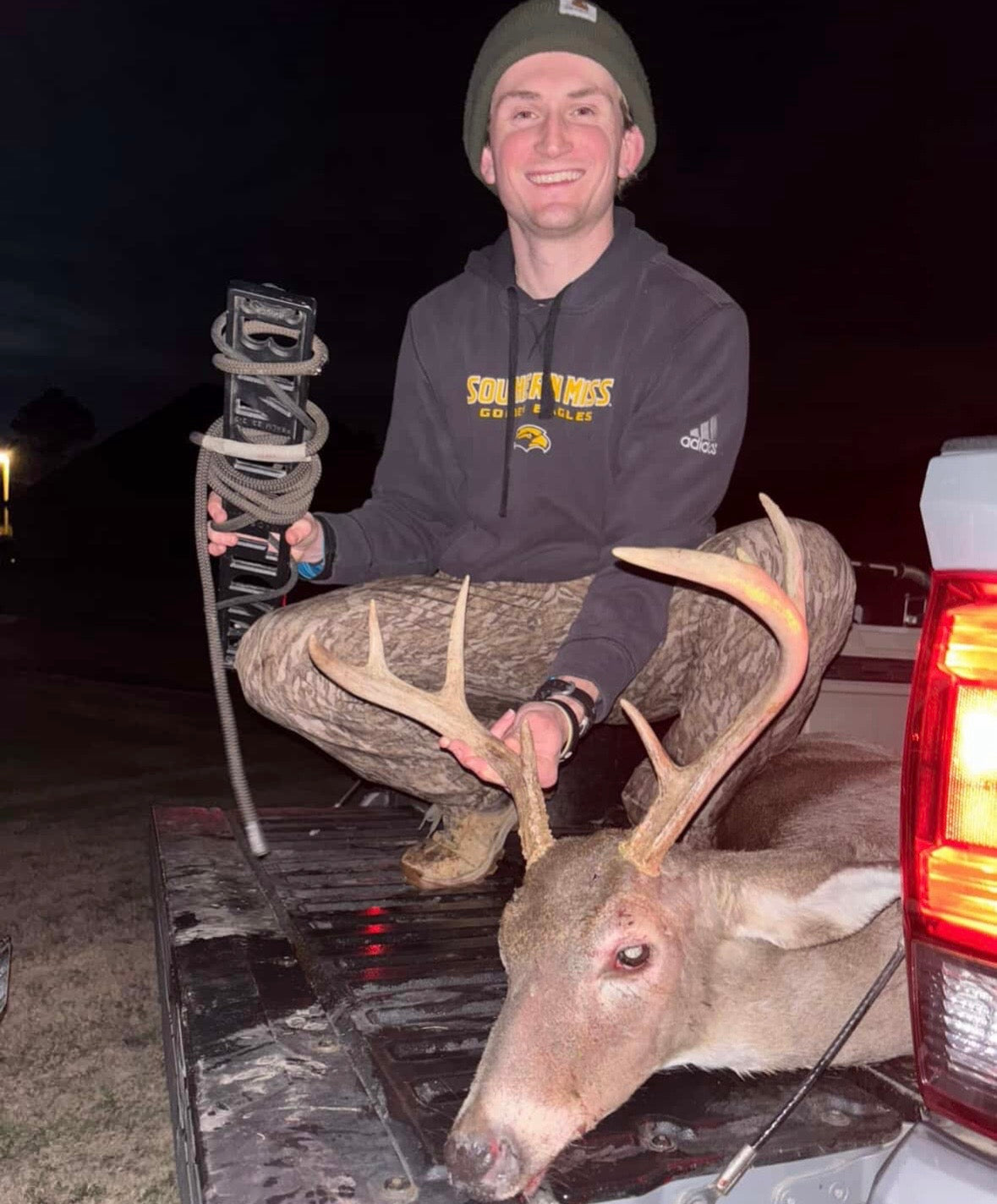 A man crouching in the bed of a pickup truck at night, holding a mounted deer head and a camera rig. He is wearing a dark gray hoodie with 'SOUTH WARD'S GREAT LAKES' and an Adidas logo, a green beanie, and camouflage pants. He is using a BANDIT Climber from Raccoon Rigs.