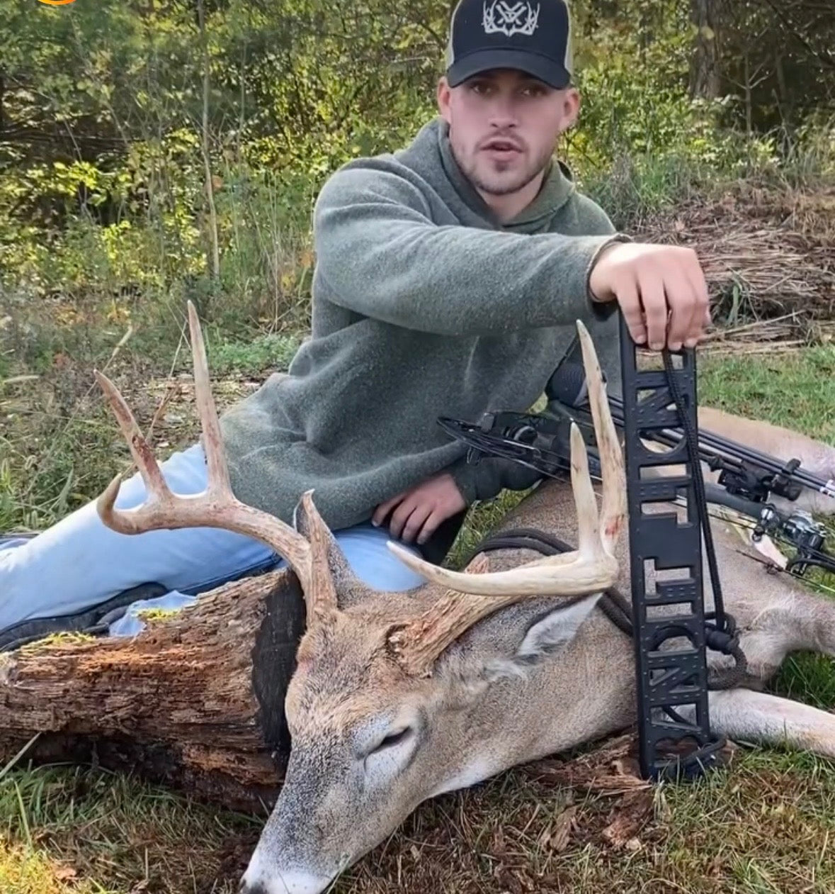 A man in a green hoodie and black cap kneels beside a harvested deer with large antlers outdoors, holding a black bow stand. A crossbow is visible on the ground in the wooded setting. He is using a BANDIT Climber from Raccoon Rigs.