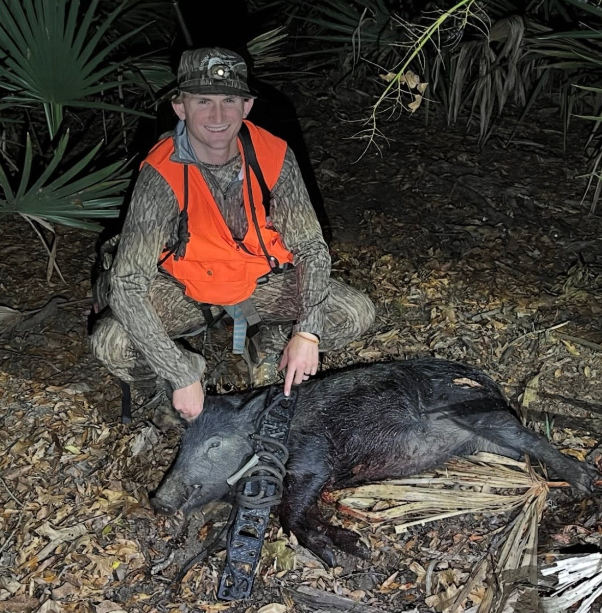 A man in camouflage hunting clothing and an orange vest kneels beside a harvested dark wild boar on leaf-covered ground at night. He is using a BANDIT Climber from Raccoon Rigs.