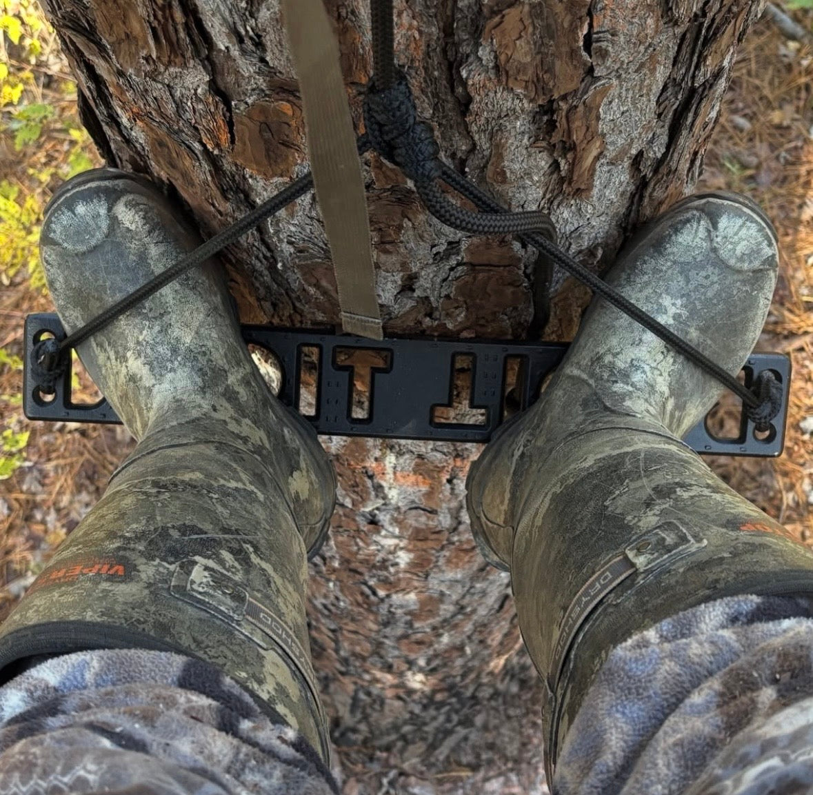 A person wearing camouflage hunting boots stands on a black metal tree ladder attached to a tree trunk in a forest setting. He is using a BANDIT Climber from Raccoon Rigs.